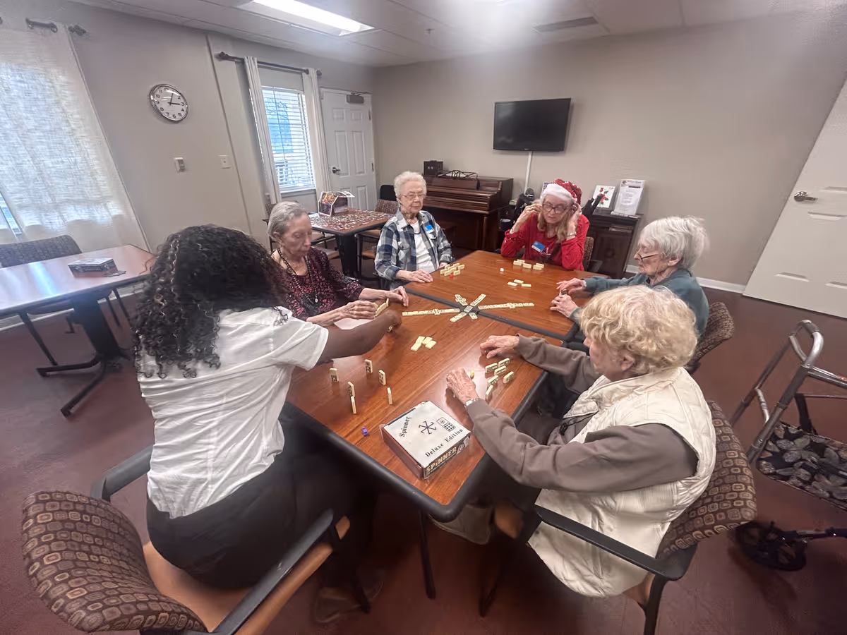 A group of elderly women and a caregiver sitting around a wooden table playing a tile-based game in a well-lit room with beige walls, a clock on the wall, a piano, and a television mounted on the wall.