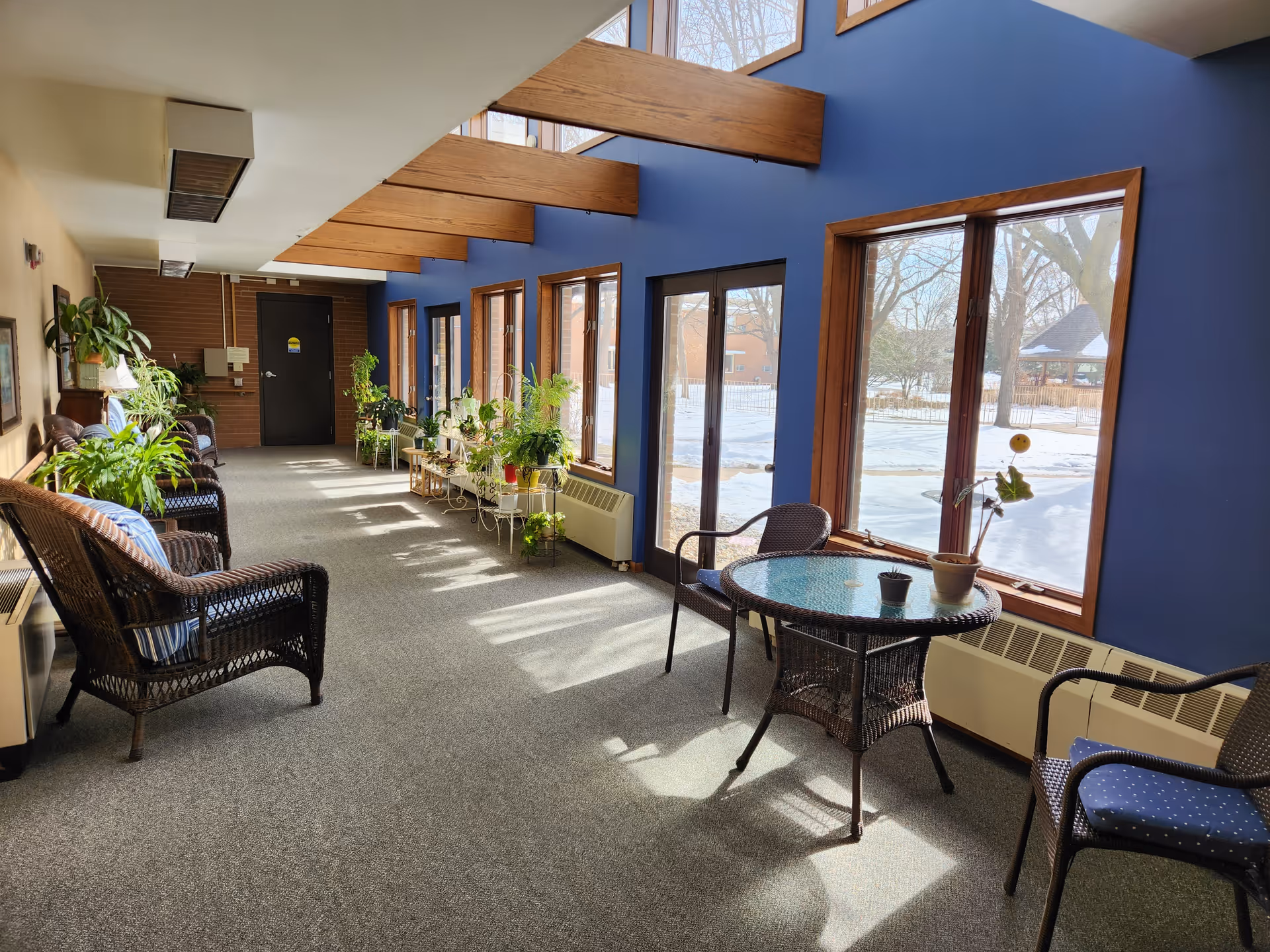 A bright indoor sitting area with large windows and glass doors letting in natural light. The walls are painted blue with wooden beams on the ceiling. There are several wicker chairs with cushions, a round glass-top table with potted plants, and multiple green plants on stands along the windows. Outside, snow covers the ground and trees are visible.