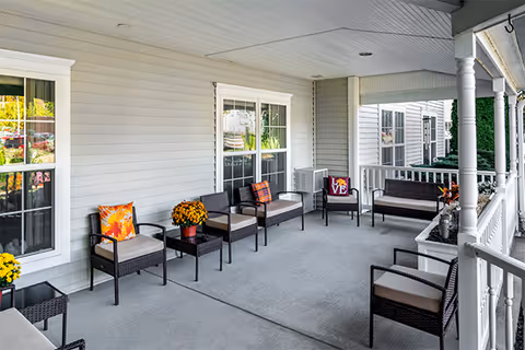 A covered outdoor porch area with multiple cushioned wicker chairs and small tables. The porch has white railings and columns, with decorative pillows and potted flowers adding color. The exterior wall of the building is light gray with white trim around the windows.