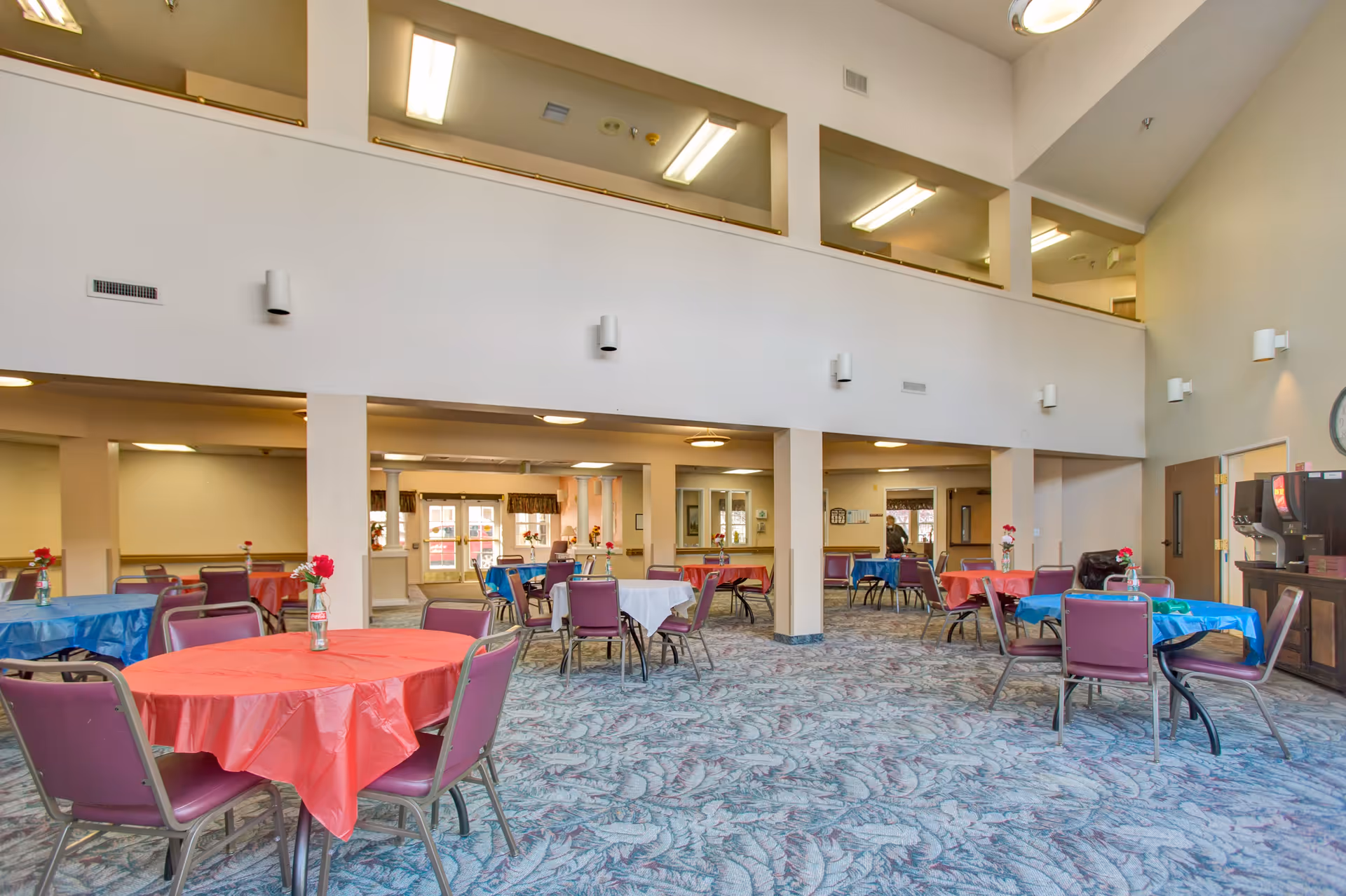 A spacious dining room in a senior living facility with multiple round tables covered in red, blue, and white tablecloths. Each table has several purple chairs around it and a small vase with a red flower as a centerpiece. The room has high ceilings with a second-floor balcony overlooking the area, patterned carpet flooring, and bright overhead lighting. A beverage station is visible on the right side of the room.