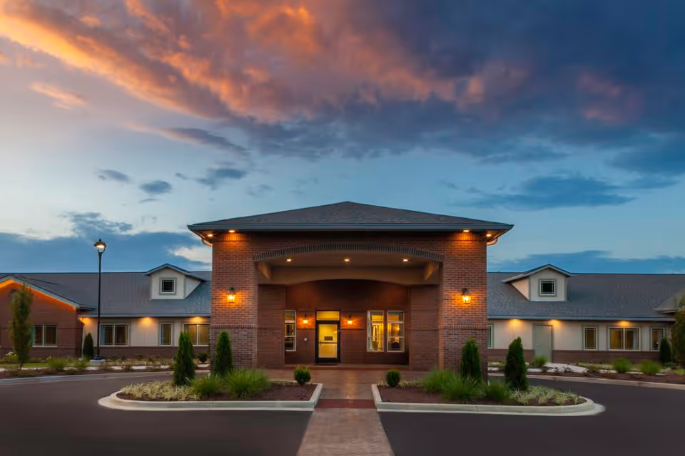 Exterior front view of Whitetail Springs Memory Care Community building at dusk, showing a brick entrance with warm lights and a landscaped driveway under a dramatic sky with orange and blue clouds.