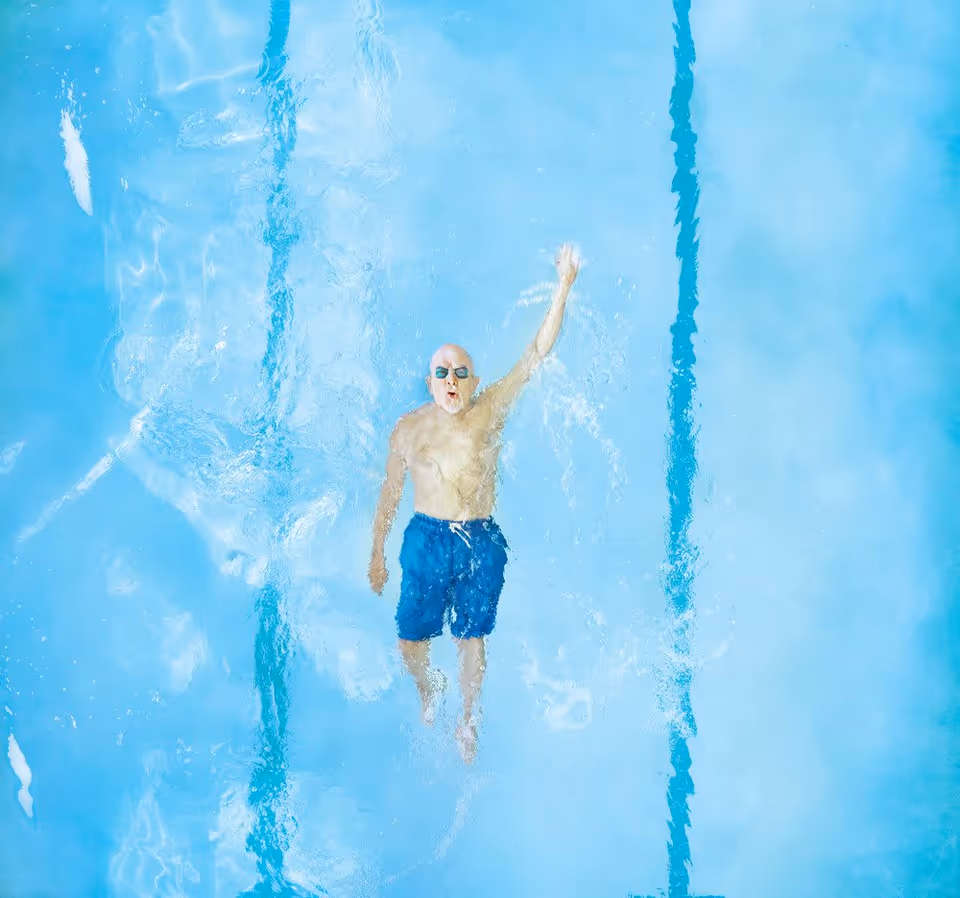 An elderly man wearing blue swim trunks and goggles swimming in a pool, viewed from above with water ripples around him.