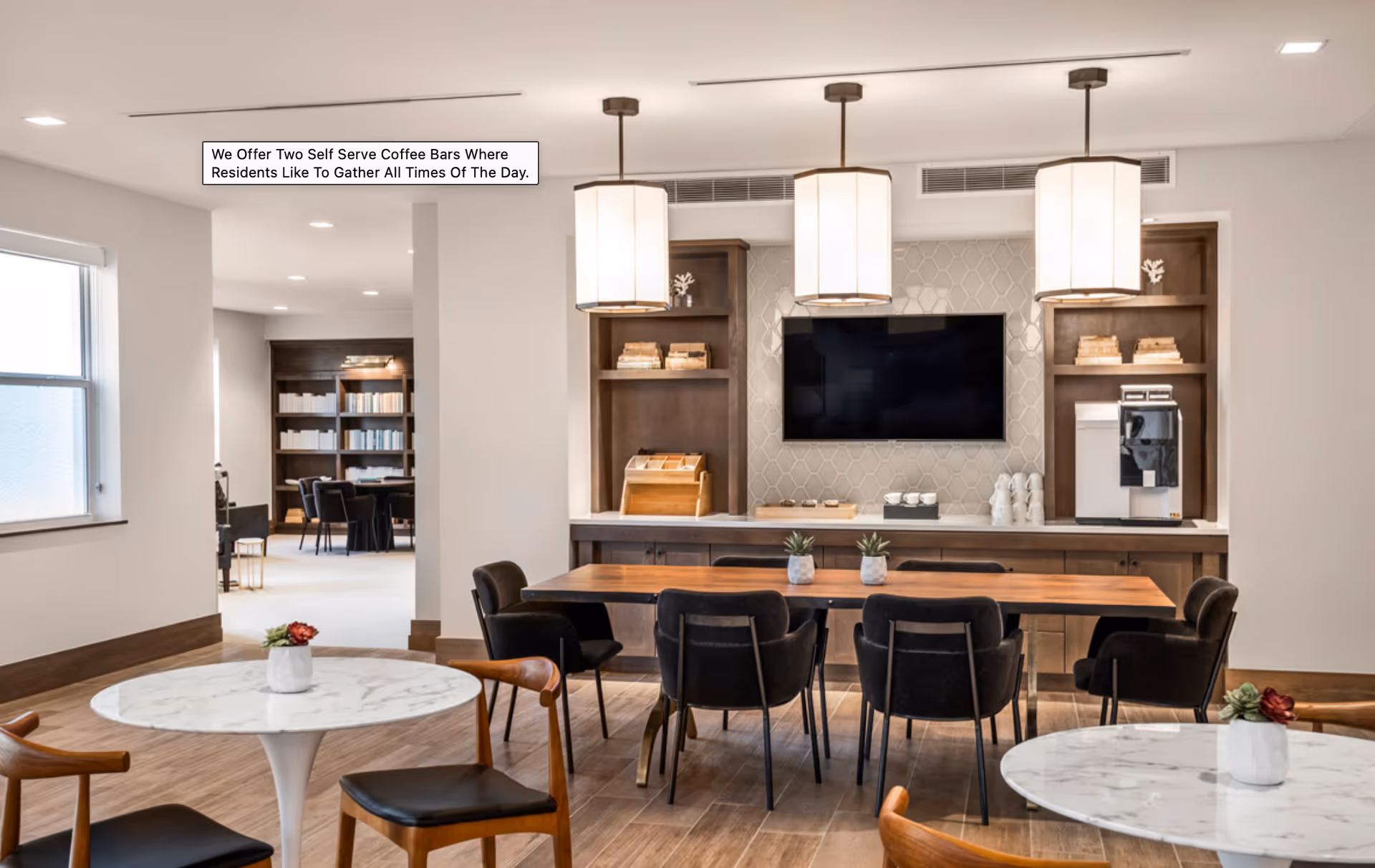 A modern communal area in a senior living facility featuring a wooden table with black chairs, two round marble tables with wooden chairs, and a self-serve coffee bar with a coffee machine, cups, and shelves. The room is well-lit with three pendant lights and has a TV mounted on a tiled wall. In the background, there is a bookshelf and additional seating.