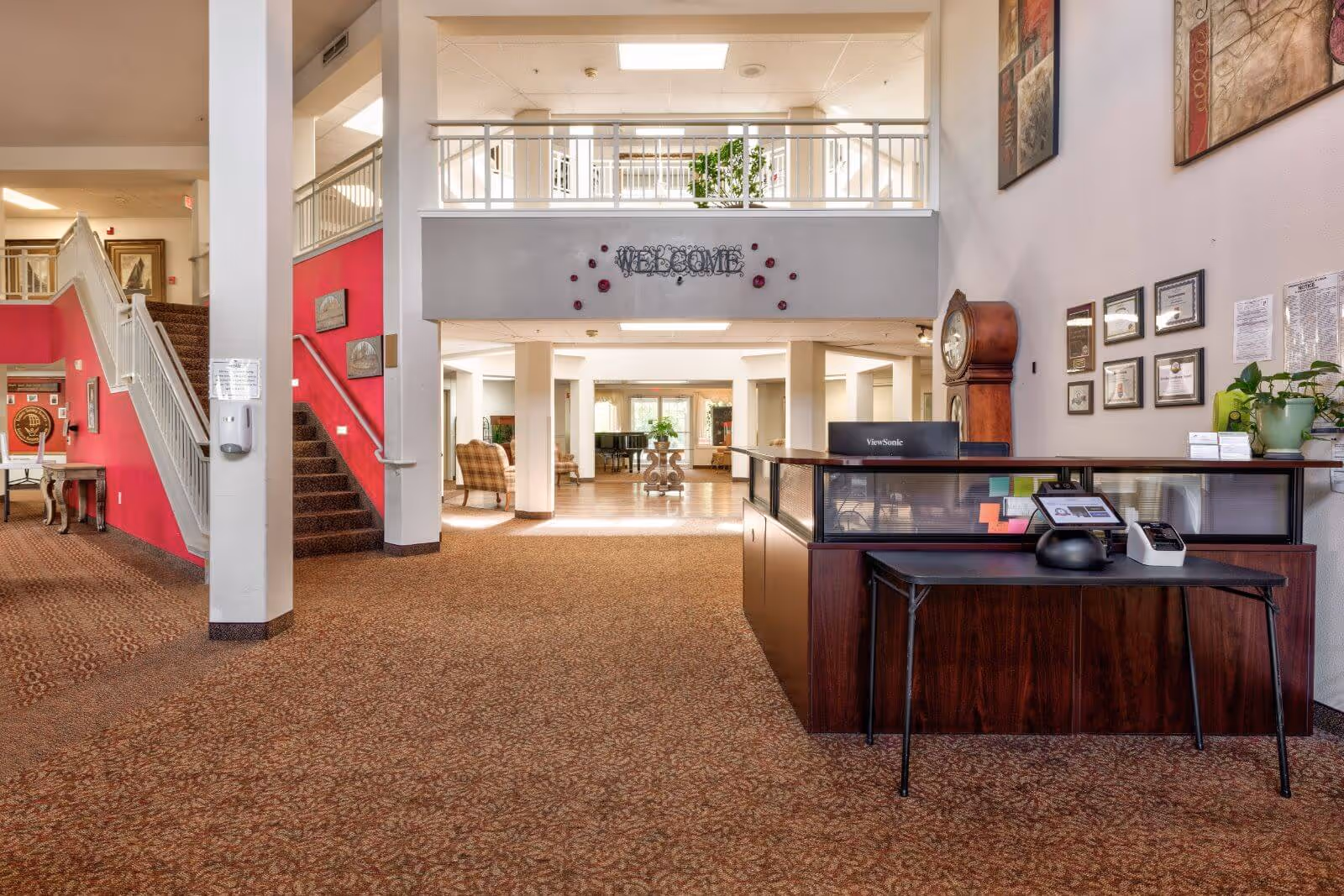 Spacious senior living facility lobby with a reception desk, seating area, staircase, and a 'Welcome' sign.