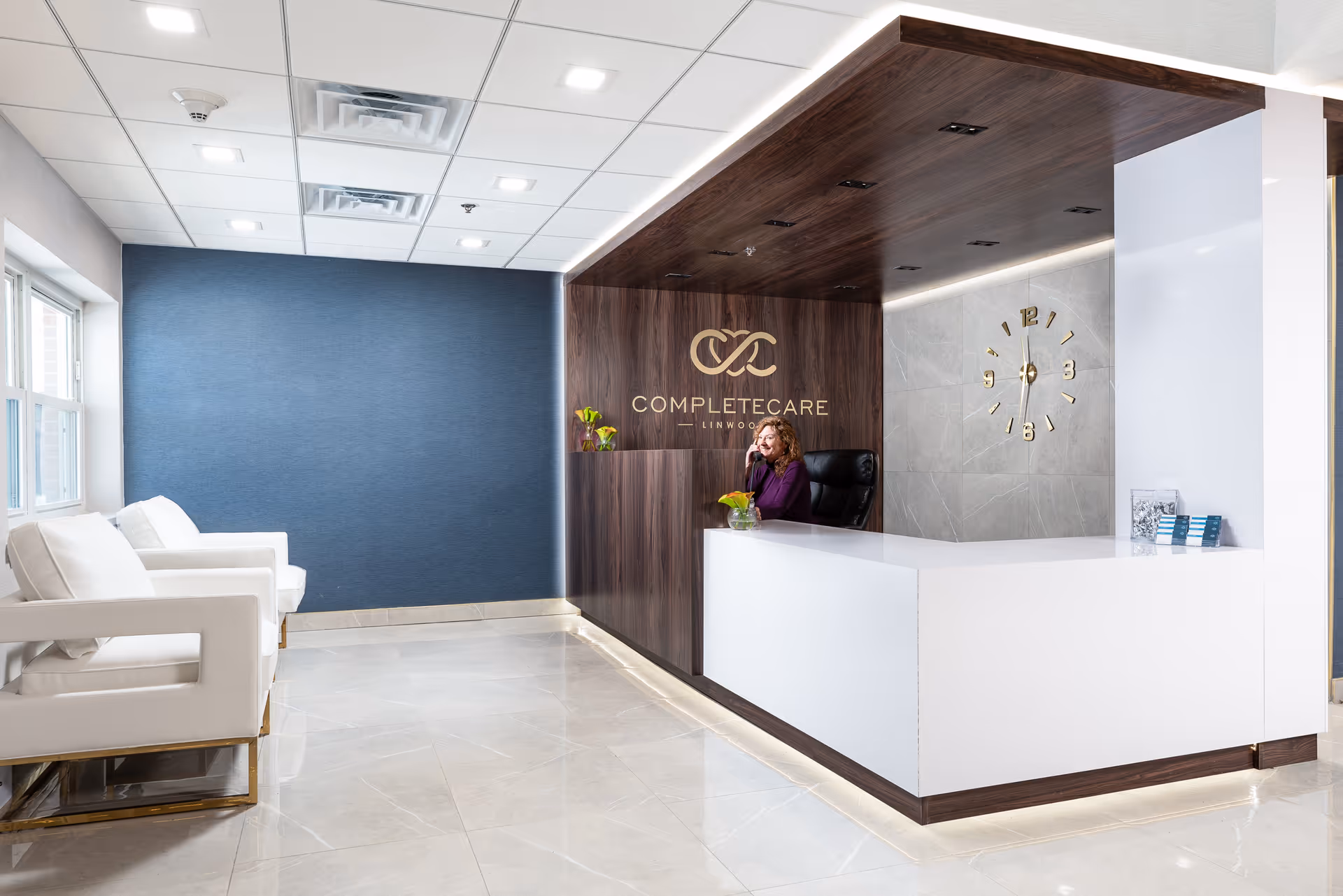 Reception area of Complete Care at Linwood with a woman sitting behind a white and wood-paneled reception desk. The wall behind her features the facility's logo and name, and a modern gold clock is mounted on a gray tiled wall. There are white armchairs along a blue accent wall on the left side of the room, and large windows letting in natural light.