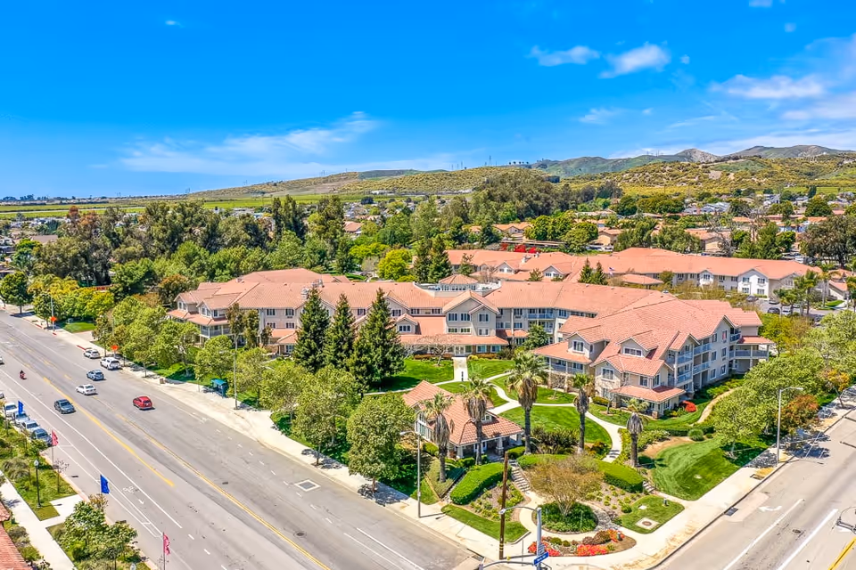 Aerial view of The Palms At Bonaventure Assisted Living & Memory Care facility showing multiple connected buildings with red-tiled roofs surrounded by green lawns, trees, and landscaped gardens. The facility is bordered by two roads with cars and street signs visible. Hills and a clear blue sky are in the background.