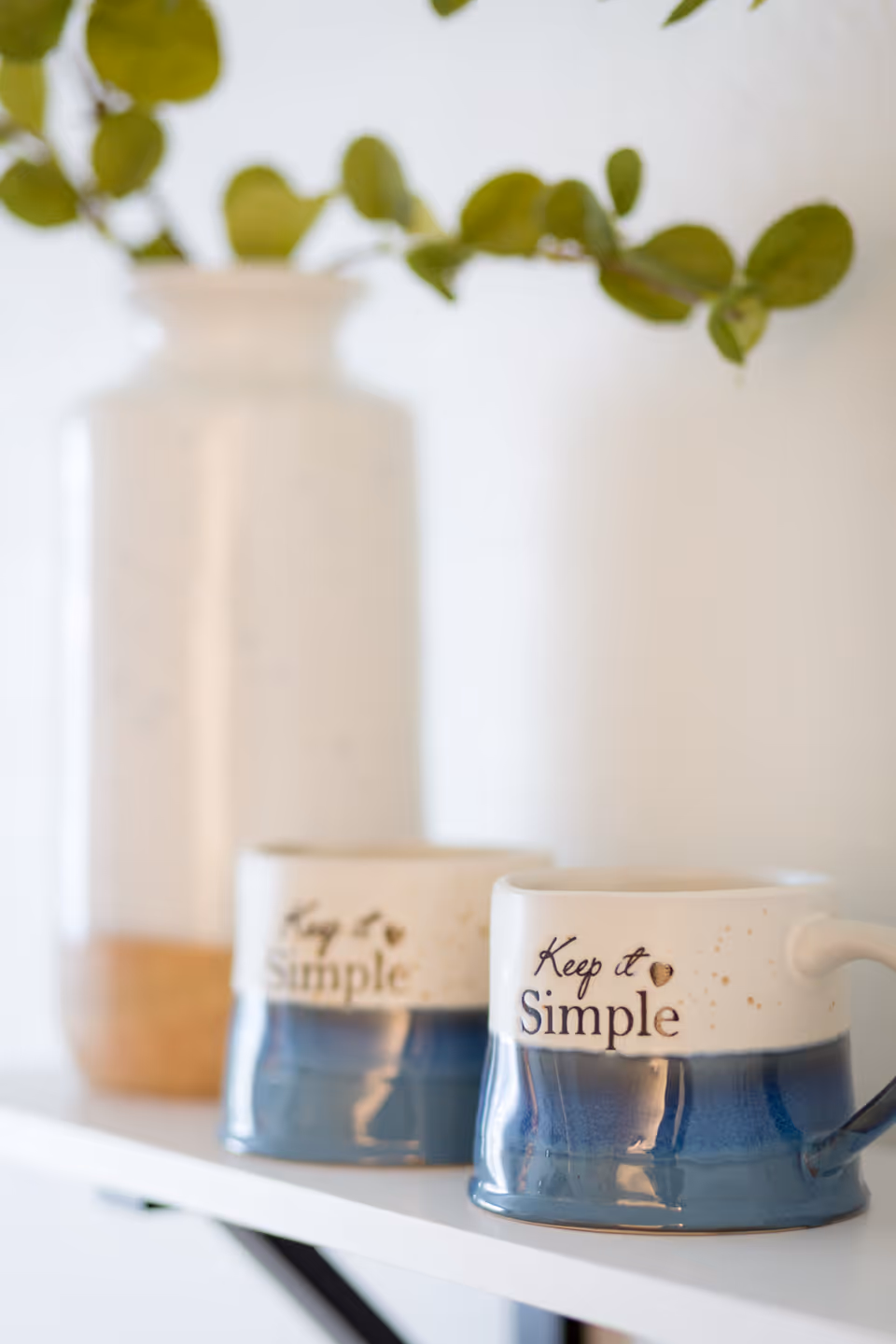 Two ceramic mugs with the phrase 'Keep it Simple' printed on them, placed on a white shelf. In the background, there is a tall ceramic vase with green leafy branches.