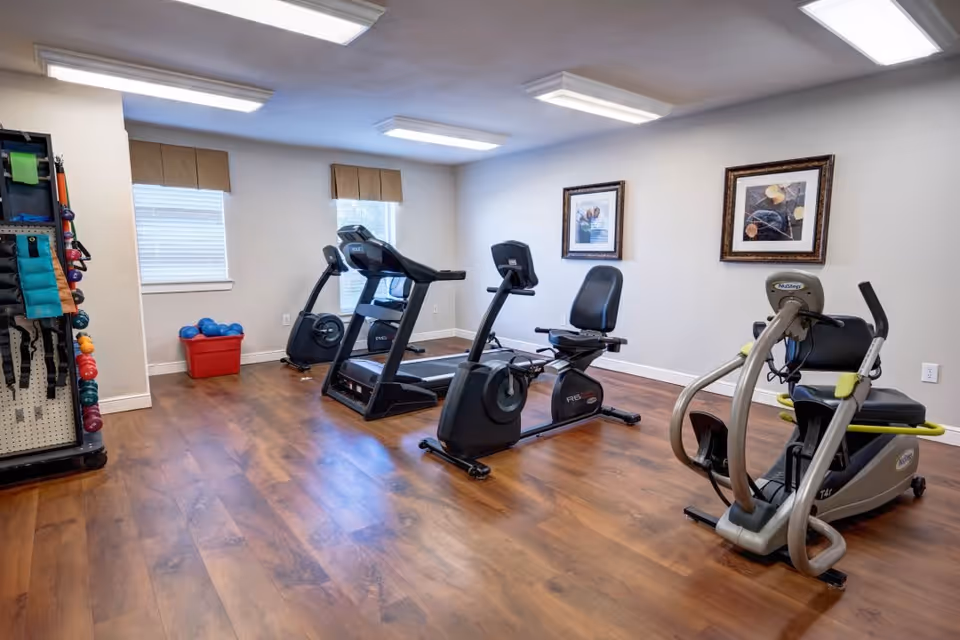 A small fitness room with wooden flooring and white walls, featuring exercise equipment including a treadmill, a recumbent bike, and a seated elliptical machine. Two windows with beige valances allow natural light into the room. On the left side, there is a rack holding various exercise accessories such as resistance bands and dumbbells. Two framed pictures hang on the wall above the exercise machines.