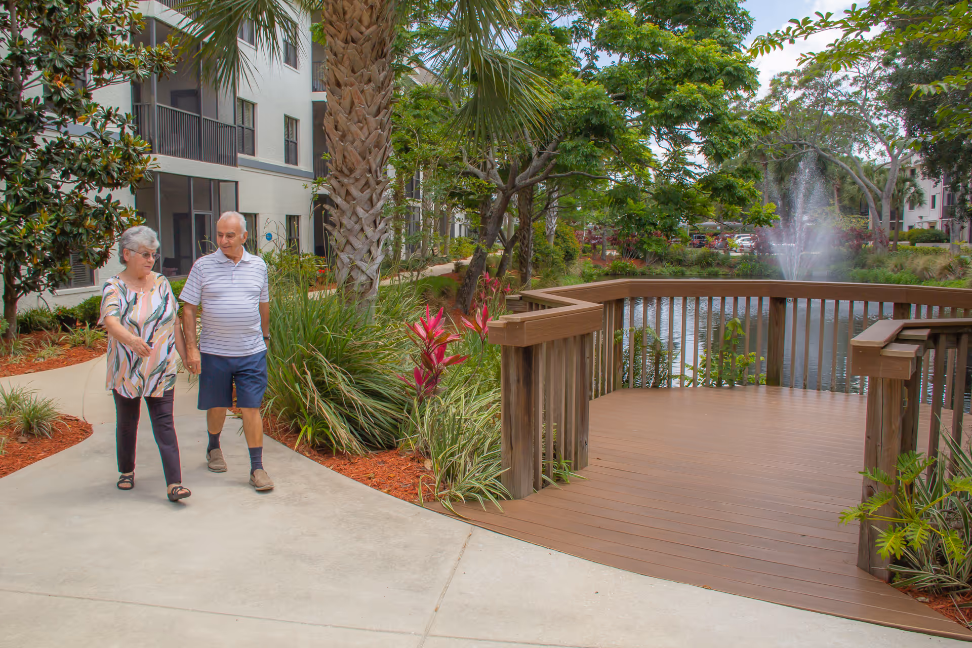 An elderly couple walking hand in hand along a paved pathway in a landscaped outdoor area of a senior living facility. The scene includes lush greenery, palm trees, colorful plants, a wooden deck overlooking a pond with a water fountain, and multi-story residential buildings in the background.