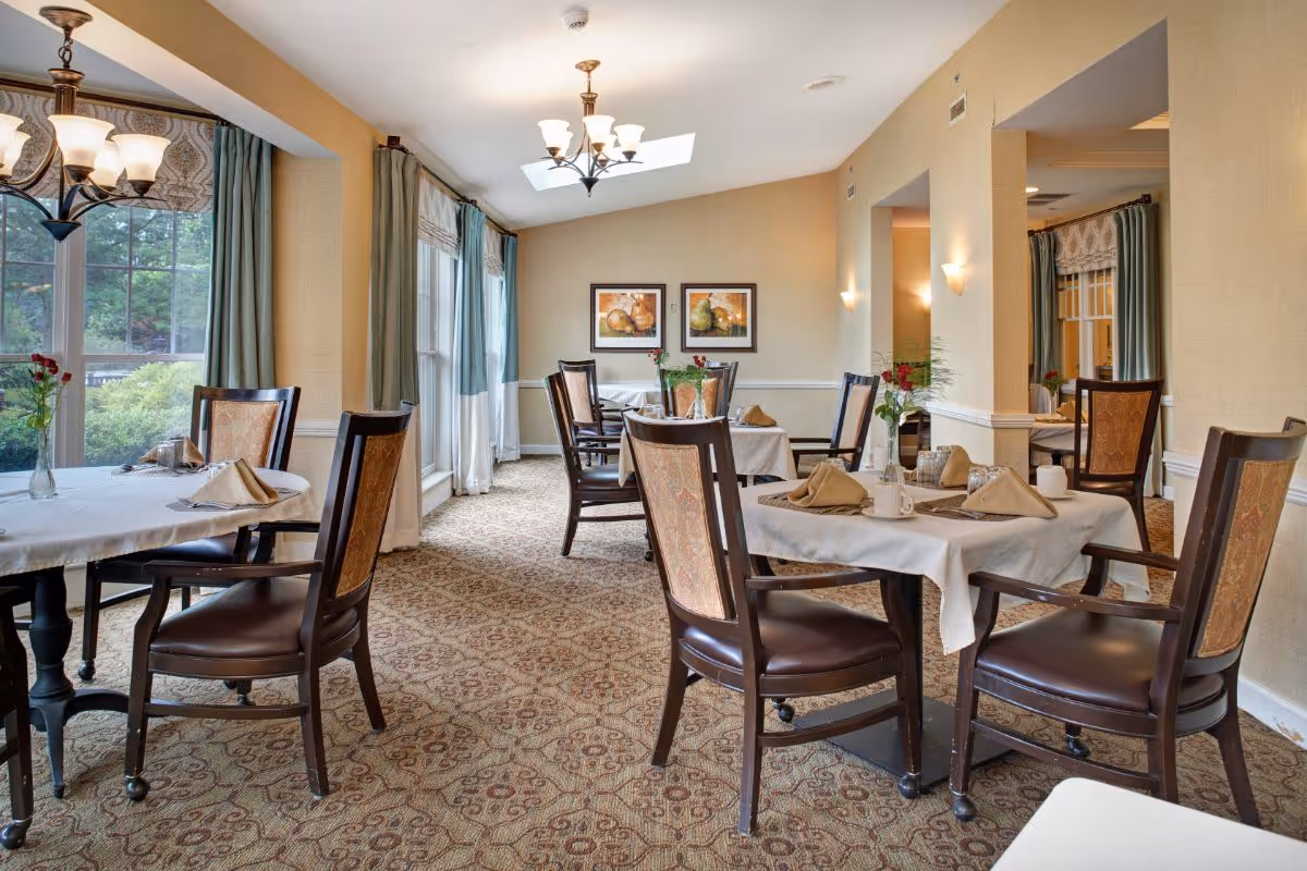 A bright dining room with several tables covered in white tablecloths, each set with napkins, glasses, and small flower vases. The room has large windows with green curtains, patterned carpet, and warm beige walls adorned with framed artwork. Elegant chandeliers hang from the ceiling providing soft lighting.