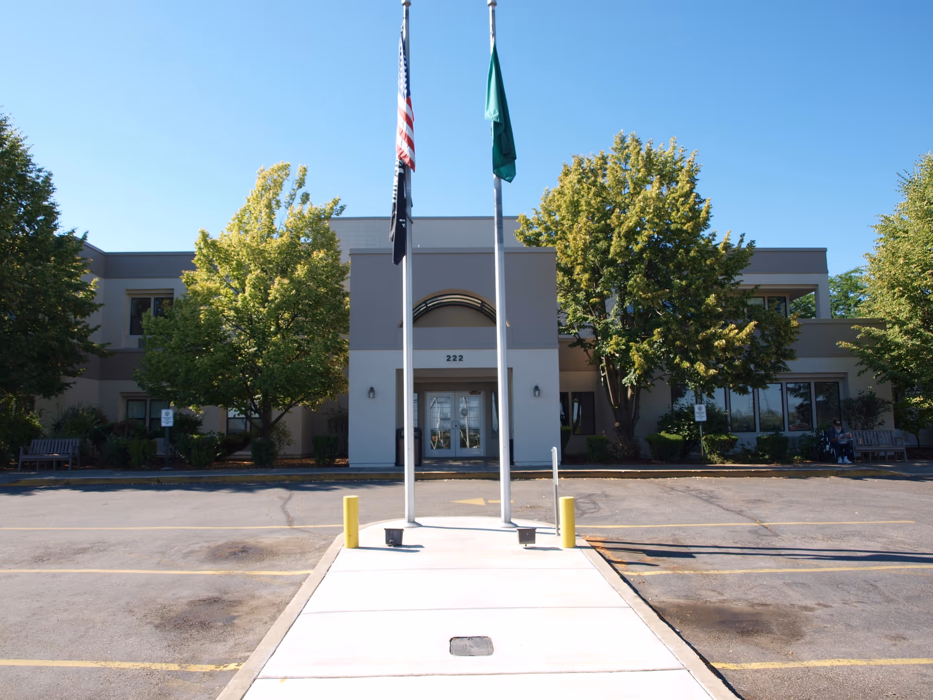 Front entrance of the Spokane Veterans Home building with flagpoles, a walkway, and trees flanking the doorway.
