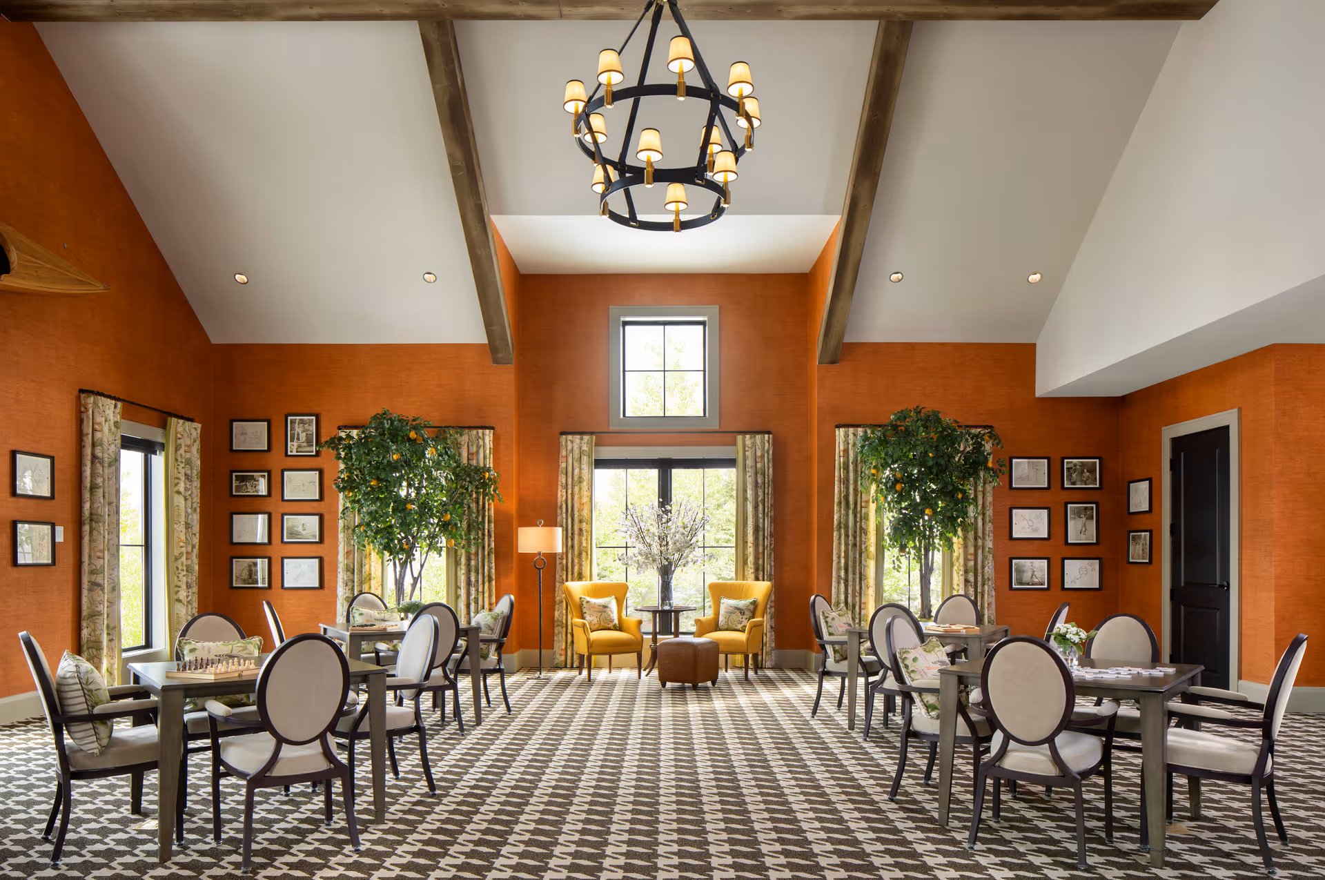A spacious and well-lit common area with high vaulted ceilings and exposed wooden beams. The walls are painted a warm orange color and adorned with framed pictures. There are multiple round tables with cushioned chairs arranged around the room, some with board games on them. Two yellow armchairs with a small round table and an ottoman are positioned near large windows with floral curtains. Two large potted plants add greenery to the space, and a large chandelier hangs from the ceiling.
