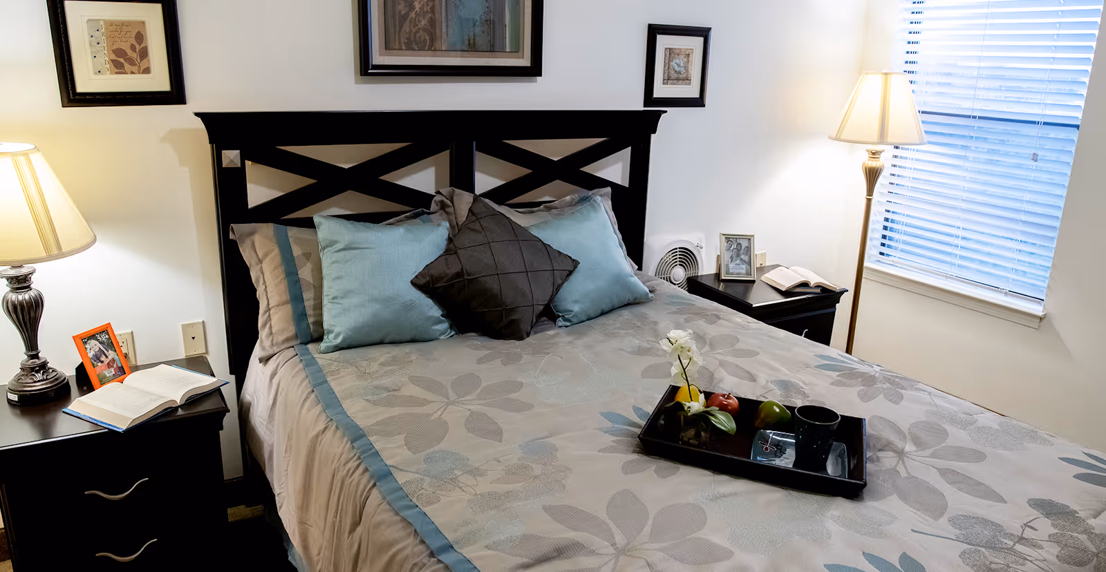 Neatly made bed with decorative pillows and a tray, flanked by nightstands and lamps in a bright bedroom.