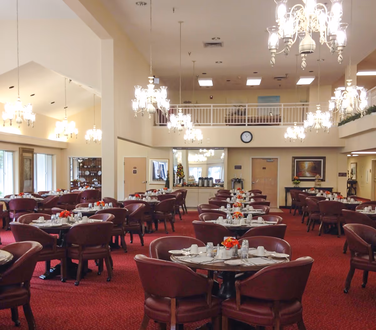 Spacious dining room with multiple round tables set with white plates, cups, and silverware. Each table has a small floral centerpiece. The room features red carpet flooring, beige walls, and several elegant chandeliers hanging from the ceiling. Large windows on the left side allow natural light to enter. A clock and framed artwork are visible on the far wall.