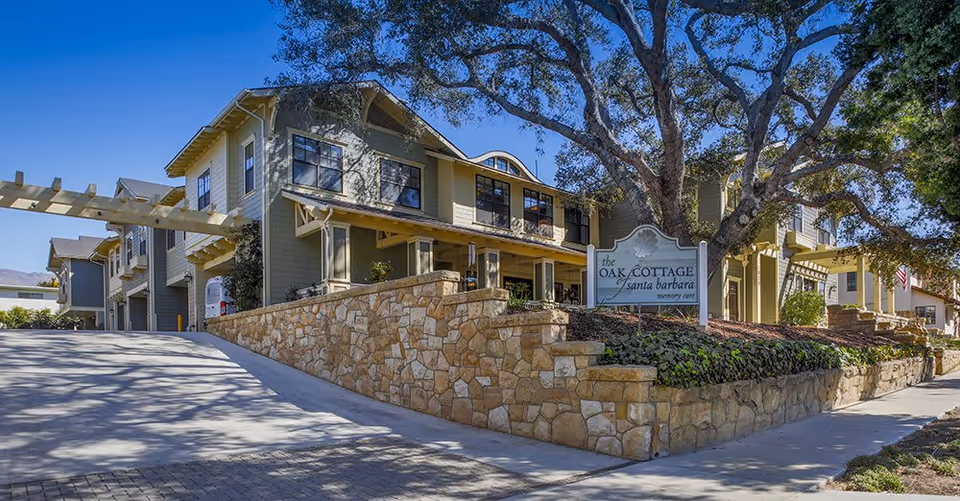 Exterior view of Oak Cottage of Santa Barbara Memory Care facility showing a multi-story building with beige siding, large windows, a stone retaining wall, and a sign with the facility's name. There are large trees providing shade and a clear blue sky above.