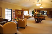 Spacious, well-lit common lounge with upholstered chairs, tables topped with floral arrangements, and a reception area in the background.