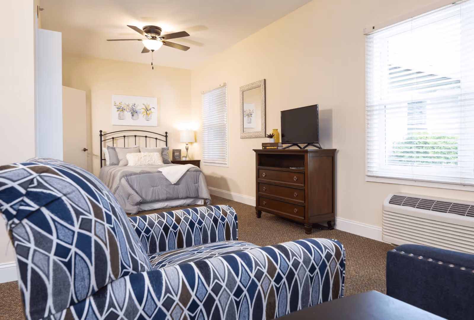 Sunny assisted-living bedroom with a bed, patterned armchair, dresser with TV, ceiling fan, and two windows.