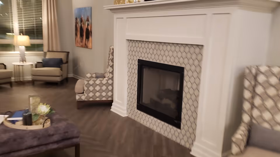 A cozy living room area featuring a white fireplace with a patterned tile surround. There are two patterned armchairs with cushions on either side of the fireplace. In the background, there are additional armchairs, a side table with a lamp, and a window with blinds and curtains. A decorative tray with a flower and other items is placed on a purple ottoman in the foreground.