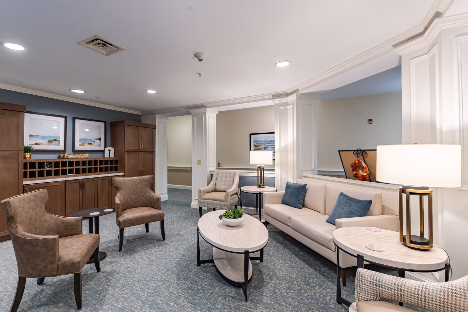 A well-lit senior living facility common area with a beige sofa, two blue cushions, two brown armchairs, a beige armchair, two round wooden side tables with lamps, a wooden coffee table with a small plant, and wooden cabinets with cubbyholes and framed artwork on the wall.