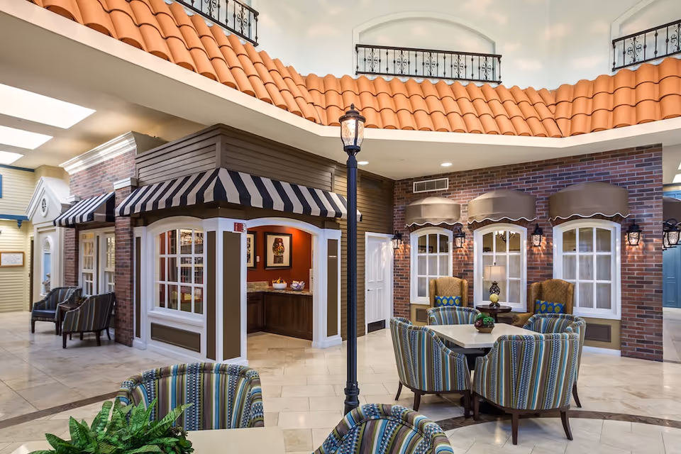 Interior common area with café-style storefronts, round tables and striped armchairs beneath an indoor tiled roof.