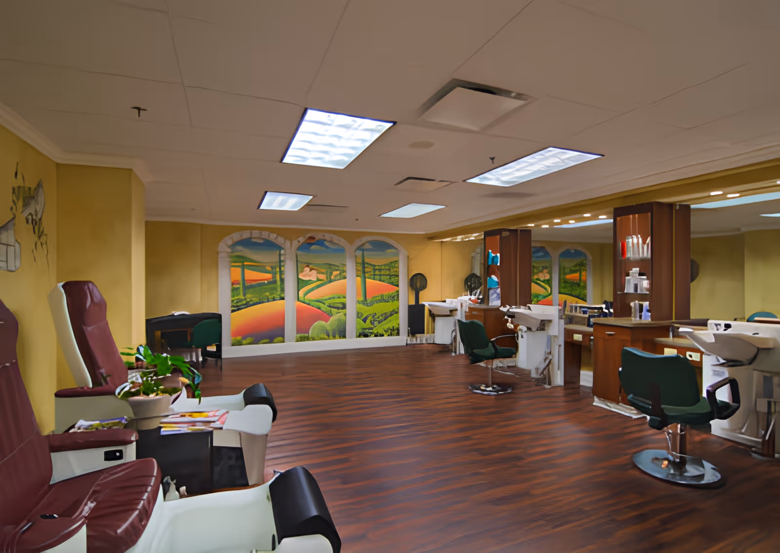 Interior of a salon area in a senior living facility with maroon and white pedicure chairs on the left, green salon chairs in front of mirrors and sinks on the right, wood flooring, yellow walls, and a mural depicting a colorful landscape with hills and fields on the far wall.