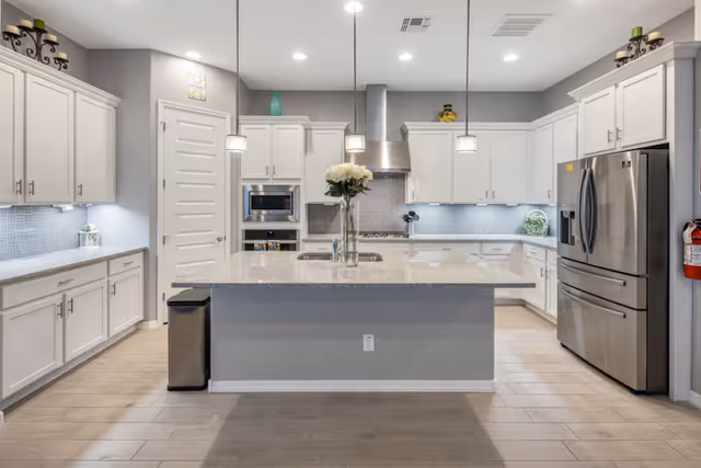 Modern kitchen with white cabinets, stainless steel refrigerator, built-in microwave, and oven. A large island with a sink and a vase of white flowers is centered under three pendant lights. The kitchen has light wood flooring and recessed ceiling lights.
