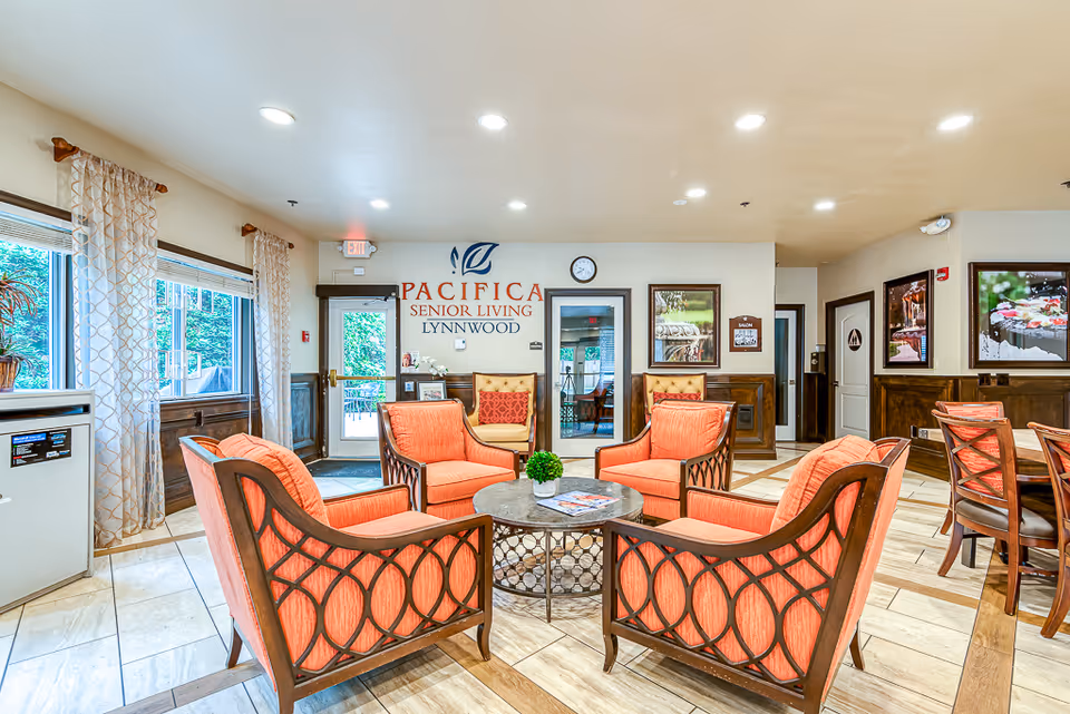 A bright and welcoming senior living facility common area with four coral-colored cushioned chairs arranged around a round coffee table with a small green plant and magazines. The room has large windows with patterned curtains, wooden paneling on the walls, and a tiled floor. On the far wall, there is a door with an exit sign above it and a sign that reads 'Pacifica Senior Living Lynnwood'. There are framed pictures on the walls and a clock above the sign.