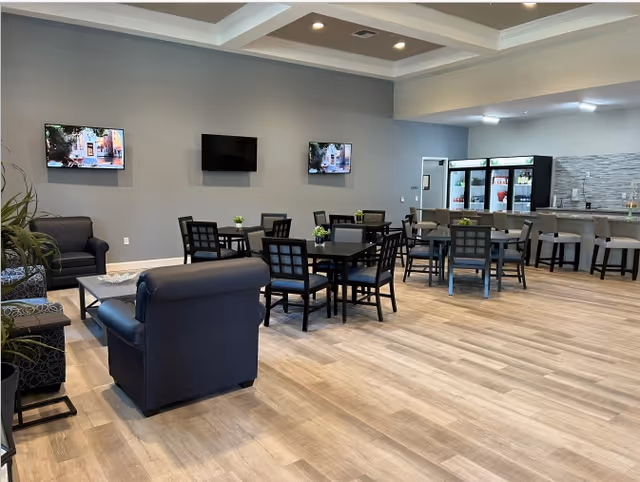 A spacious senior living common area with multiple black tables and chairs arranged for seating. There are three wall-mounted flat-screen TVs on a gray wall. The room features light wood flooring, a seating area with dark blue armchairs and a coffee table, and a counter area with bar stools and refrigerators stocked with beverages.