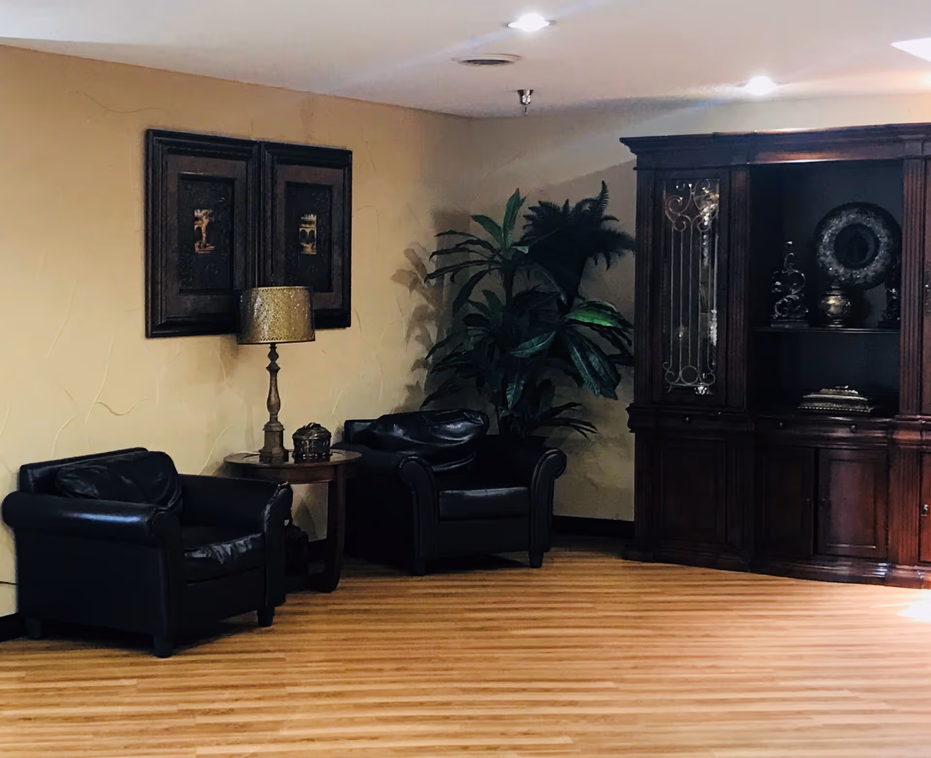 A cozy interior corner with two black leather armchairs separated by a small round wooden table holding a decorative lamp and a small container. Behind the chairs is a large green potted plant. On the wall above the table are two framed artworks. To the right is a large dark wooden cabinet with glass doors displaying decorative items. The floor is light wood and the walls are beige.