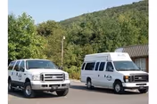 Two white vehicles parked on a paved area in front of a building with a brown roof, surrounded by trees and a green hillside in the background.