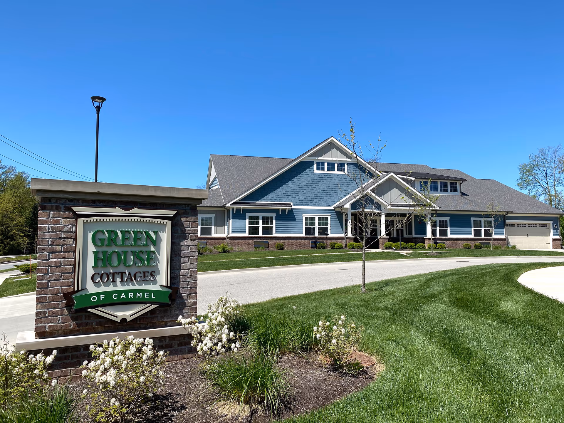 Blue cottage-style senior living building with a brick sign reading "Green House Cottages of Carmel" and landscaped lawn under a clear blue sky.