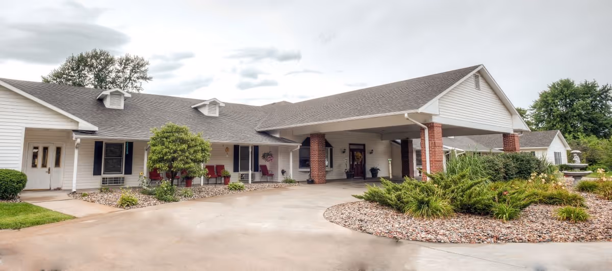 Exterior view of a single-story senior living facility building with white siding and a gray roof. The entrance has a covered driveway supported by brick columns. There are shrubs, small trees, and landscaped rock beds around the building, with a concrete driveway and walkway leading to the entrance.