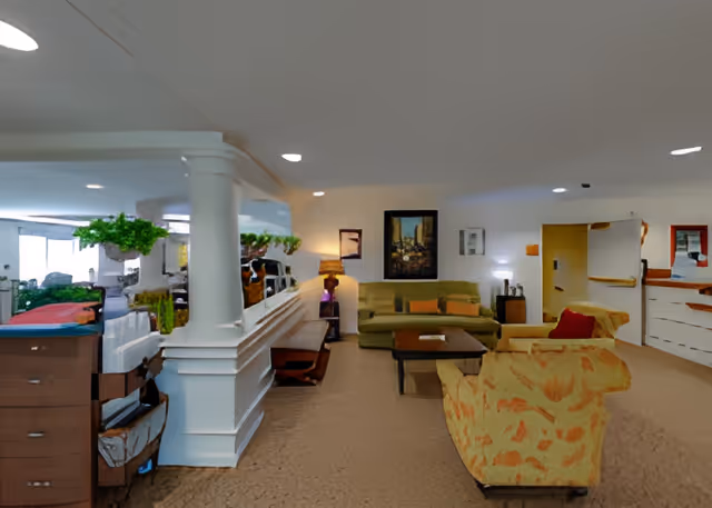 Interior view of a living room area in a senior living facility with beige carpet, a green sofa, a yellow patterned armchair, a wooden coffee table, and a side table with a lamp. There are framed pictures on the walls and a large mirror reflecting part of the room.