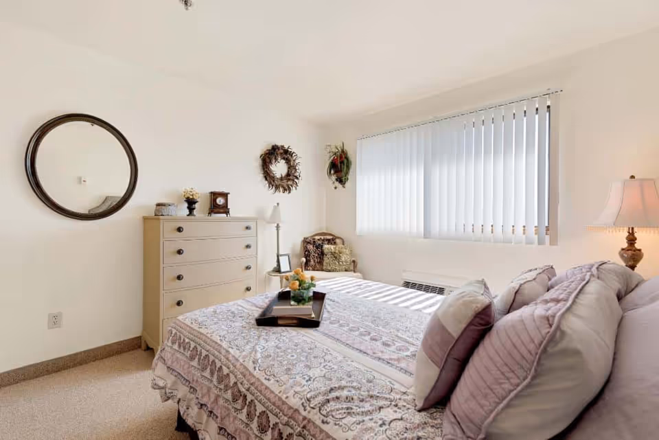 Bright, softly decorated bedroom with a made bed, dresser topped with decor, round wall mirror, vertical blinds over a window, and a bedside lamp.