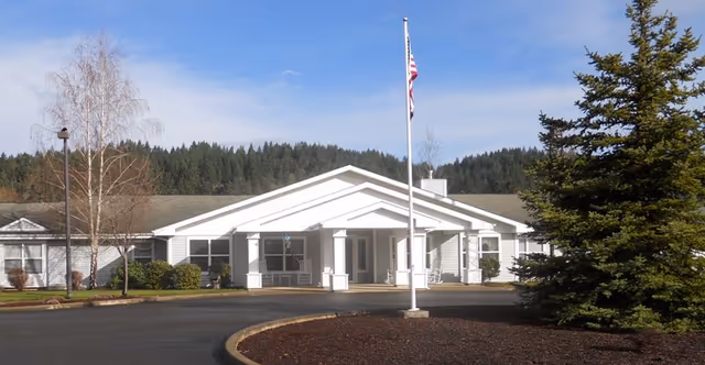 Exterior view of a single-story senior living facility building with a white entrance porch, an American flag on a flagpole in front, surrounded by trees and landscaping under a clear blue sky.