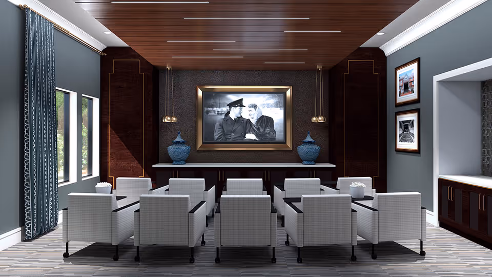 A small theater-style room with two rows of white cushioned chairs facing a large framed black and white movie screen. The room has dark wood paneling on the back wall, blue-gray walls, two blue decorative vases on a console table beneath the screen, and patterned curtains on the left side windows. Two framed pictures hang on the right wall.