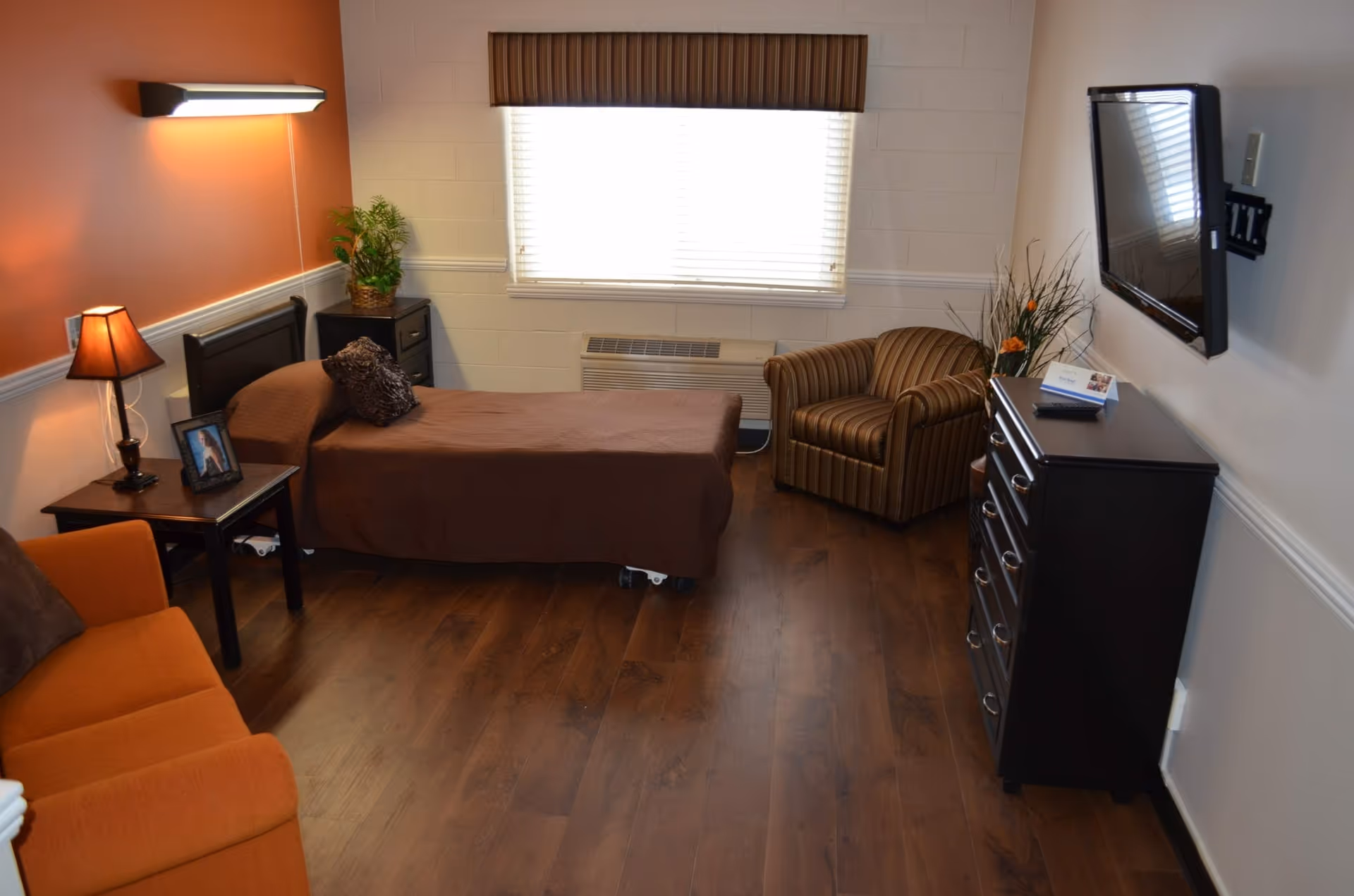 Furnished senior resident room with a single bed, sofa and armchair, dresser with a wall-mounted TV, and a window.