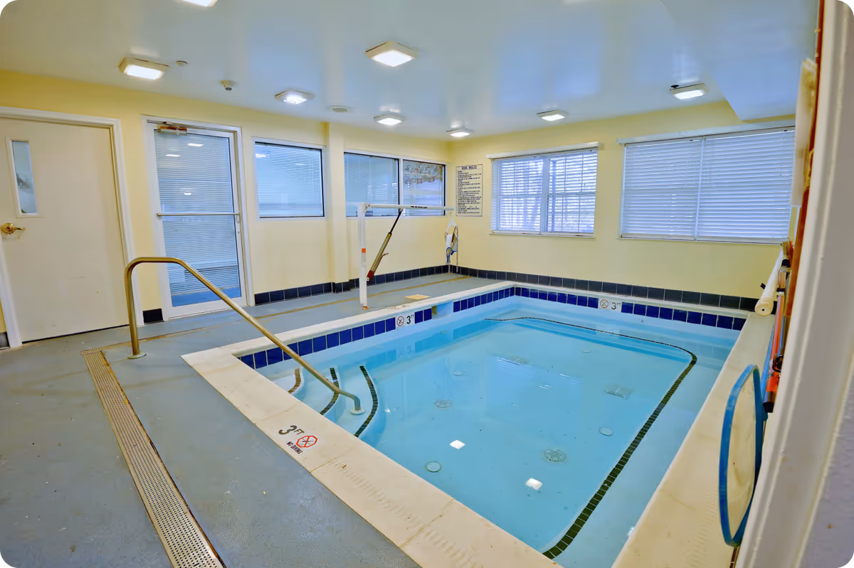 Indoor small swimming pool with handrails and a pool lift for accessibility, surrounded by yellow walls and windows with blinds. The pool area has a non-slip floor and ceiling lights.