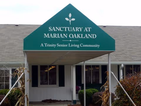 Entrance canopy with green triangular sign reading 'Sanctuary at Marian Oakland, A Trinity Senior Living Community' above a covered walkway leading to the building entrance, surrounded by shrubs and plants.