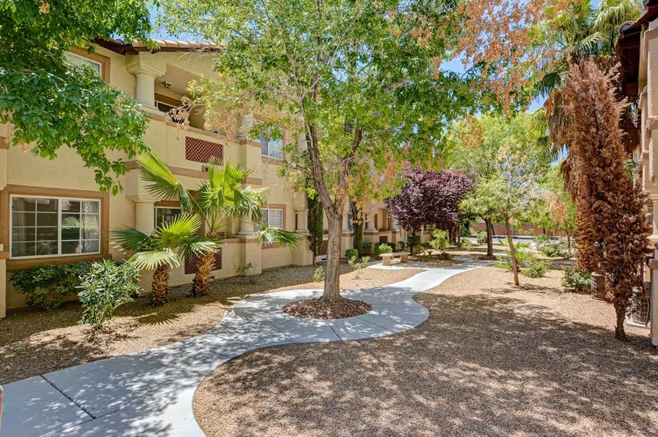 Outdoor courtyard area at Oakey Assisted Living featuring a winding concrete pathway surrounded by trees, shrubs, and desert landscaping with beige two-story buildings in the background.