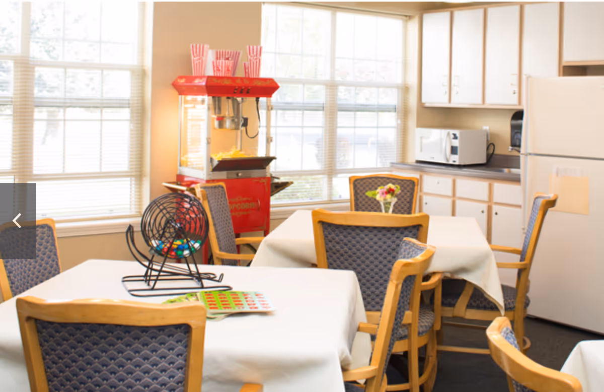 A bright room with several tables covered in white tablecloths and surrounded by wooden chairs with blue patterned cushions. A red popcorn machine is positioned near large windows letting in natural light. On one table, there is a bingo cage with colorful balls and bingo cards. The room also features a white refrigerator, microwave, and cabinets along one wall.