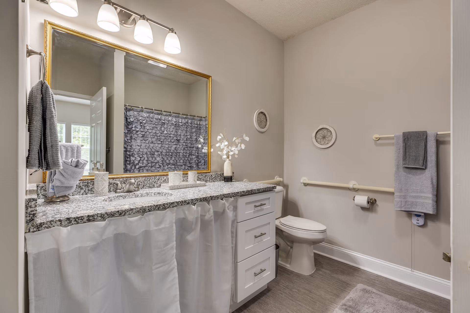 A clean and modern bathroom with a large granite countertop and white cabinetry. There is a large mirror with a gold frame above the sink, three light fixtures above the mirror, and a gray towel hanging on a rack. The bathroom has a toilet with grab bars on the wall, a shower with a gray patterned curtain, and decorative wall hangings. The floor is covered with gray tiles and a matching bath mat.
