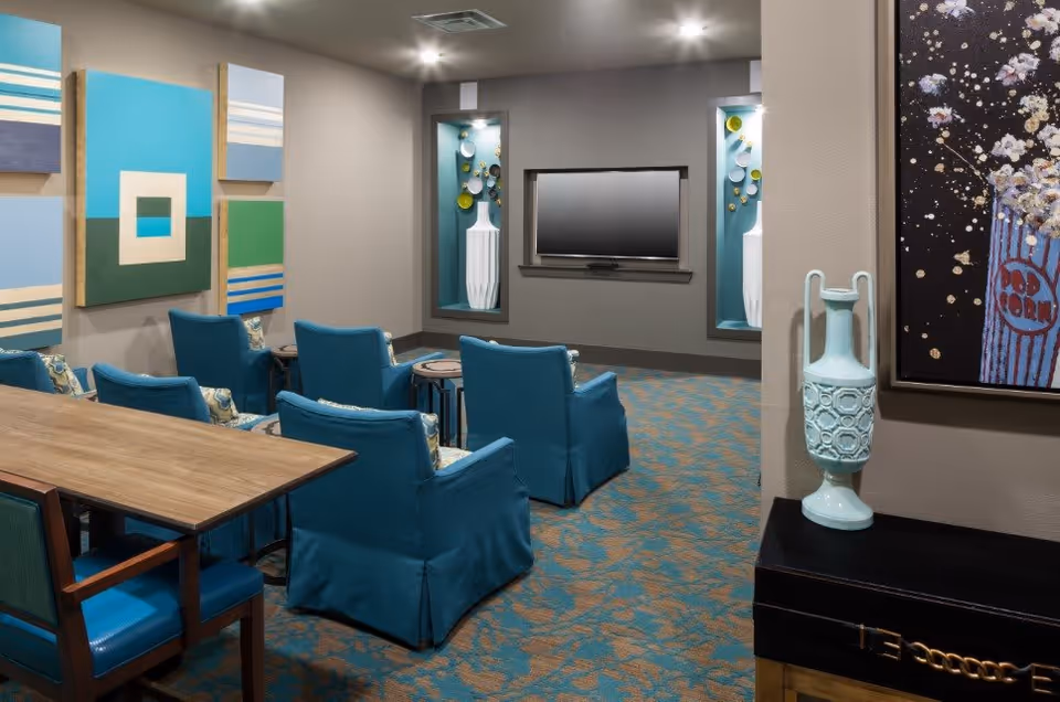 A cozy living room area with six blue upholstered chairs arranged in two rows facing a wall-mounted flat screen TV. The room features a patterned carpet in blue and beige tones, modern abstract wall art, and decorative vases displayed in recessed wall niches. A wooden table with blue chairs is partially visible in the foreground.