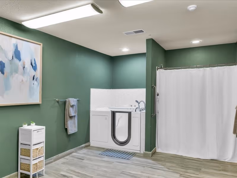 Accessible bathroom with green walls featuring a walk-in tub with a U-shaped door, a shower with white curtain, towel rack, and framed artwork.