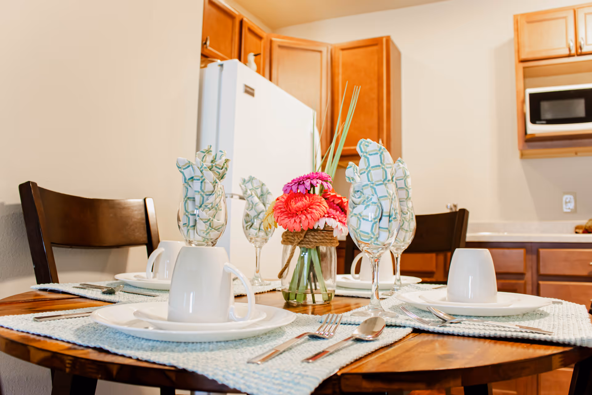 A wooden dining table set for four with white plates, cups, silverware, and glasses with patterned napkins inside. A small vase with pink and red flowers is in the center of the table. The background shows a kitchen area with wooden cabinets, a white refrigerator, and a microwave.