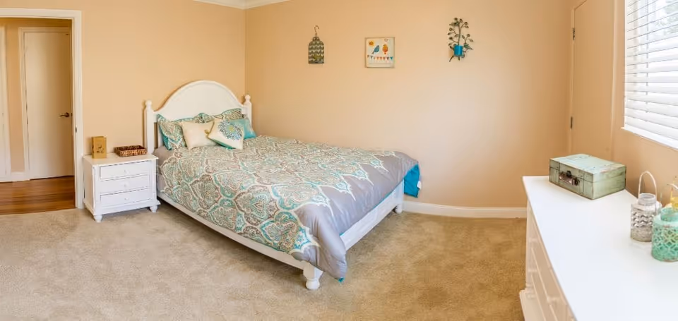 A cozy bedroom with beige walls and carpeted floor. The room features a white bed frame with a patterned blue and gray bedspread and several pillows. There is a white nightstand with a small basket and decorative item next to the bed. On the opposite side, a white dresser holds decorative jars and a small green box. The room has a window with white blinds and three small wall decorations above the bed.