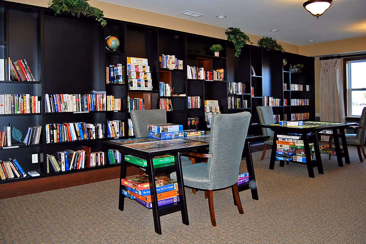 Community library/activity room with dark bookshelves, tables and chairs, and stacks of board games and puzzles.