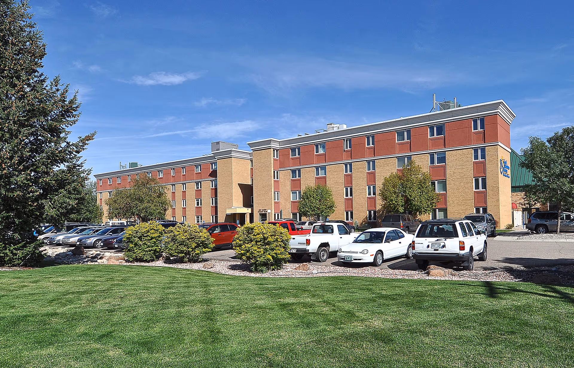 Exterior view of a multi-story senior living facility building with a parking lot in front. Several cars are parked, and there are green bushes and trees around the building. The sky is clear and blue.