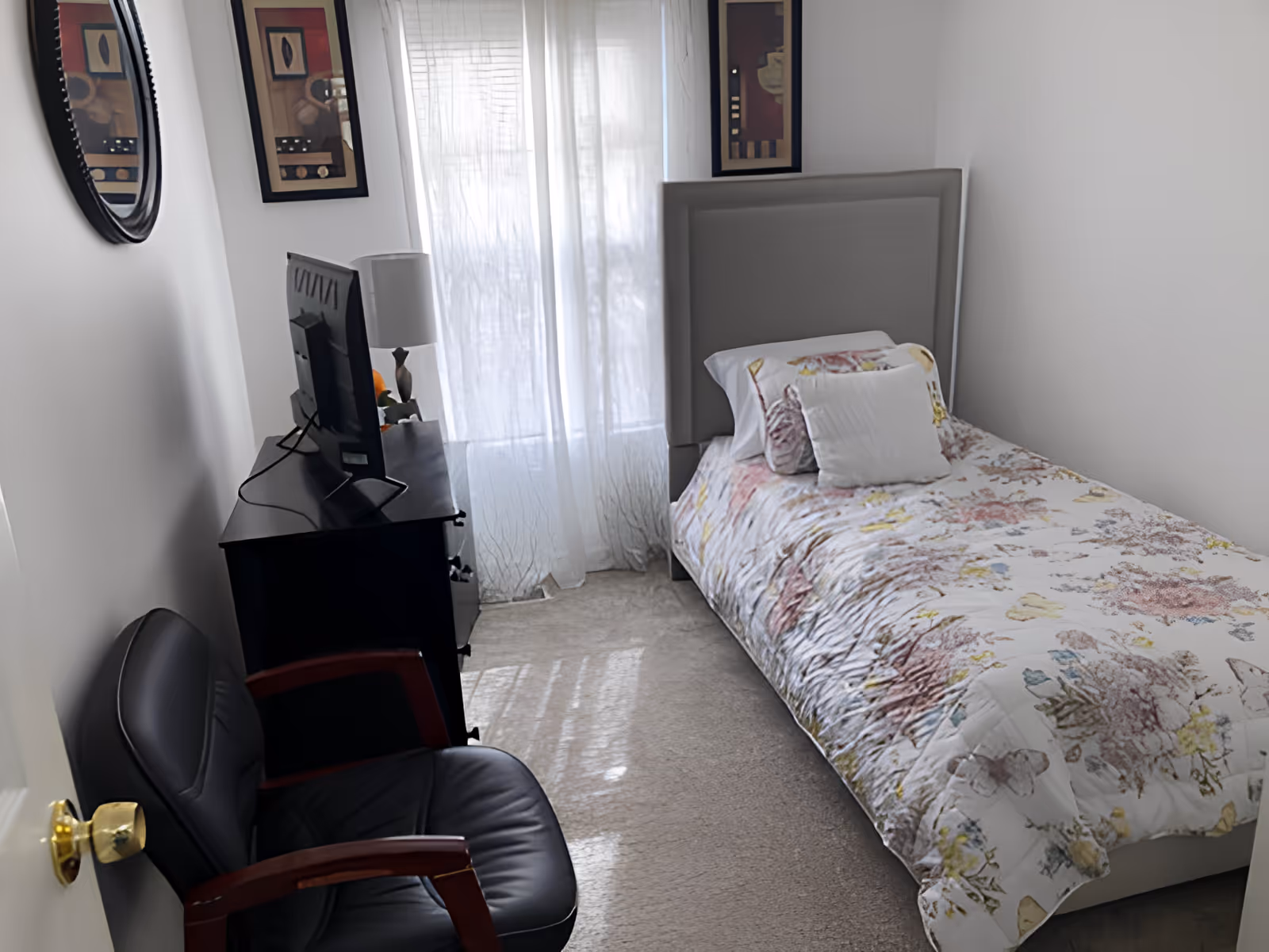 Small single bedroom with a twin bed covered in a floral comforter, a dresser with a TV, an armchair, and a window with sheer curtains.