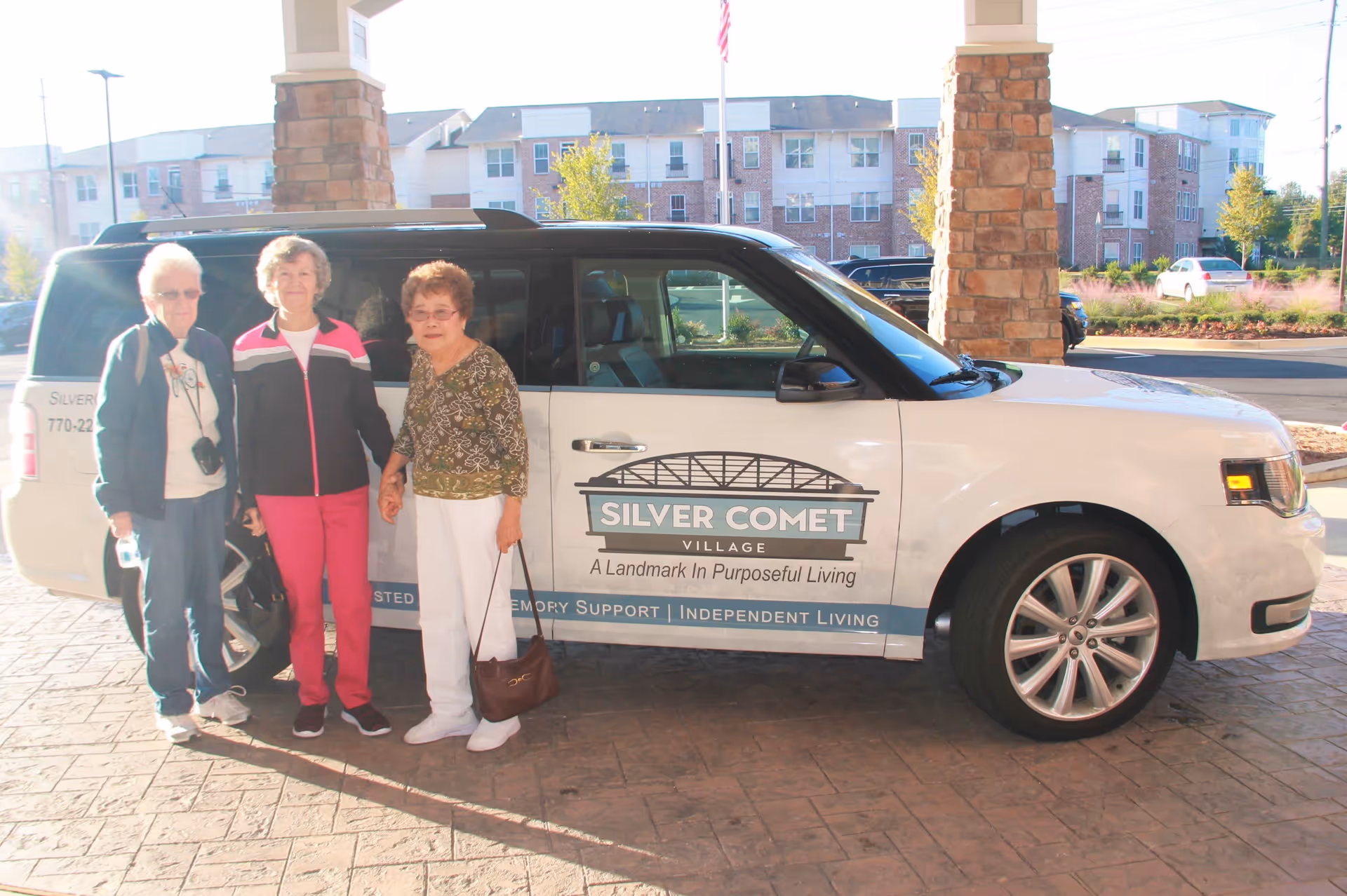 Three elderly women standing next to a white vehicle with Silver Comet Village branding parked under a covered entrance. The vehicle has text indicating services like assisted living, memory support, and independent living. Residential buildings and landscaping are visible in the background.